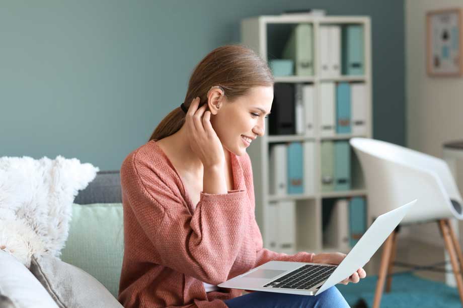 Young woman siting on a bed with hearing aid using laptop